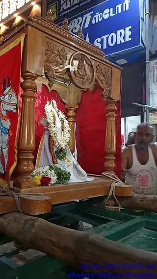 A photograph of a wooden palanquin-style vahana shrine on a green wheeled wooden platform in a busy street. Inside the carved wooden canopy, a deity murti in white robes with a white and green flower garland and a loose flower offering at its base sits in front of a red curtain. A red cloth panel to the left shows a painted bull with a decorated harness. A bare-chested man in a white vest stands beside the shrine, and a blue shop sign is visible above. A timestamp reads 28 Dec 2025 8:08:42 pm.