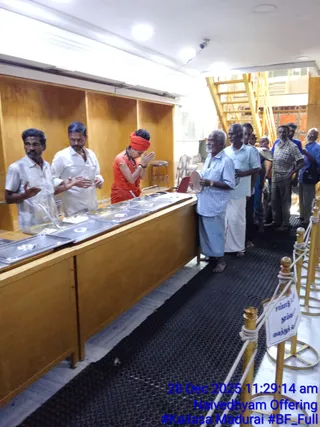 Indoor photo of a food serving counter with steel trays of prepared food under bright ceiling lights. Three people stand behind the wooden counter: two men in white shirts and white trousers on the left, one with hands pressed in anjali, and a woman in an orange sari-style top with an orange head wrap in the centre, hands joined in prayer. On the right, a line of older men and others waits, one elderly man in a light blue checked shirt and blue dhoti holds a steel plate. A yellow metal scaffold and a gold rope barrier with a Tamil sign are visible. Overlaid blue timestamp reads 28 Dec 2025 11:29:14 am, Naivedhyam Offering.