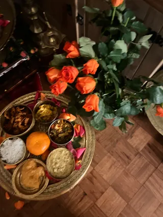 Photograph of a food offering on a round brass thali tray set on a wooden parquet floor. The tray holds small brass bowls of rice, curried dishes, a whole orange, fried bread and a bowl of pale sauce, scattered with pink and orange rose petals. Beside it stands a large bouquet of orange roses with green leaves in a vase. Brass ritual vessels and lamp stands are visible in the dim background. No people are visible.
