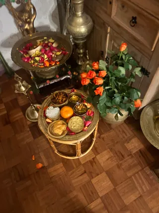 Indoor photo of a devotional offering arrangement on a parquet wooden floor. In the centre, a round rattan side table holds a brass tray with small brass bowls of cooked food, a whole orange, white rice, and scattered pink and red petals. Behind it stands a large brass tray filled with red, pink and yellow flower petals on a brass stand. A brass oil lamp with a long stem stands to the left, and a tall ornate brass vase sits near a wooden cabinet. A potted plant with orange roses is at right. No people are visible.