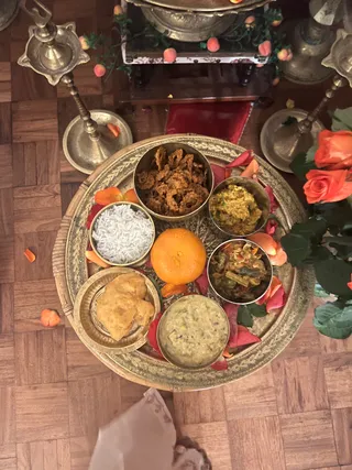 Overhead photo of a brass thali on a wooden parquet floor, set before a shrine. The round engraved brass tray holds small bowls of white rice, a fried crispy dish, a yellow-green curry, a spiced vegetable dish, a dal-like preparation, a whole orange and a puri. Scattered red and orange rose petals decorate the tray and floor. Two tall brass oil lamps stand on either side, and orange roses sit at the right edge. A shrine with peach-colored flowers and a brass urn is visible at the top. No people are visible, only a partial pale garment at the bottom edge.