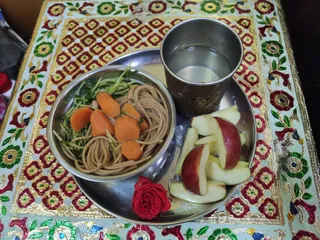 A top-down photo of a meal served on a stainless steel thali placed on an ornate table or tray with gold-edged red, green and white floral and geometric painted border. In the upper right sits a stainless steel tumbler filled with water. In the lower left, a small stainless steel bowl holds whole wheat noodles or pasta mixed with sprouted green sprouts or leafy greens and cooked orange carrot pieces. To the right on the thali are sliced red-skinned apples, and a single red rose lies at the lower edge. No people are visible.