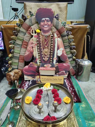 A photograph of a Pratyaksha Pada Puja setup: a life-size standing cutout photograph of a smiling man in a maroon turban with a red bindi, wearing layered gold necklaces, rudraksha-style beaded malas and a flower garland, holding a trident and seated on a patterned cushioned throne with peacock-eye style decorations. The cutout is a printed image, not a physical person. In front, a brass tray holds a pair of silver padukas (sandals) decorated with red and yellow roses and white flowers, with small brass lamps beside them. A book titled 'Living Enlightenment' sits on a red cloth at the base, next to a sign reading 'Pratyaksha Pada Puja'. A wooden elephant figurine, a brass bell, a steel vessel and a green silk cloth complete the arrangement.