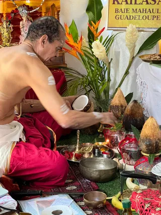 A photograph of a shirtless man with short greying hair and white ash markings on his shoulders and arm, seated on a red cloth and seen in profile. He wears a white dhoti with a red lower garment and holds half of a fresh coconut over a ritual setup. In front of him are brass and steel vessels, a red-stained plate, a lit oil lamp, a metal bowl, banana fruit, and several coconuts with leaves set on red-netted pots. Behind him stands a yellow sign reading KAILASA BAIE MAHAULT GUADELOUPE, with orange heliconia, white ginger flowers and palm fronds. A small deity murti with a decorated headdress stands on a shelf at the upper left. The scene appears to be a puja or abhishekam-style offering in a temple setting.