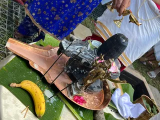 A photograph of a Shiva lingam being bathed during abhishekam at an outdoor temple setting. The dark, polished lingam sits on a copper plate-shaped basin with water flowing over it, and a small brass Nandi-type figure stands beside it with pink flower petals scattered around. A ripe banana rests on a green banana leaf on the left, with incense sticks laid across the leaf. Behind, a person in a white dhoti with gold border holds a brass vessel pouring water, and a woman in a blue floral sari stands at the left edge. Folded white cloth sits on the right near a brass pot.
