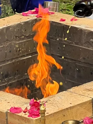 A square brick fire pit with a tall orange flame rising from its centre. Red and pink flower petals are scattered on the pit's rim and on the ground at its base, and a brass vessel sits at the bottom edge. A stream of liquid. No people are clearly visible, only a blue fabric edge at the top left. This appears to be a homa or yagna fire offering.