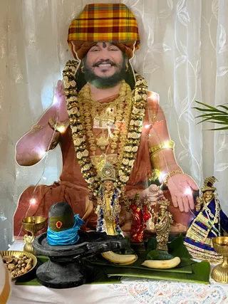 A photograph of a home shrine set against sheer white curtains with patterned leaf motifs. A large life-size cutout print shows a smiling bearded man in a plaid madras-check head wrap, orange-brown robes and a garland of white roses with gold beads, raising one hand in blessing. Before it stands a black stone shivalinga wrapped with a blue cloth and a red bindu, with bananas on banana leaves, a brass oil lamp, a small gold deity murti in red and gold clothing, and a gold ornamented figure in a blue shawl. Fairy lights are wound around the cutout and a brass bowl of rudraksha seeds sits at the left edge.