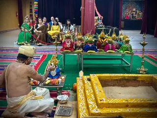 A bare-chested priest in a cream and gold-bordered dhoti and a cream turban sits cross-legged on a green and multicoloured striped mat in front of a yellow-painted square fire pit filled with sand, applying something to a row of pots. Several kalasha pots dressed in green, red, blue and pink cloth with mango-leaf and flower garlands are arranged on steel tables, with fruit and offerings on a tray beside the priest. Brass oil lamps stand on either side, and a small deity murti sits near the pit. Seated spectators in saris and salwar kameez watch from chairs behind a red rope, and a colourful deity backdrop with a peacock design hangs on the wall. The setting suggests a homa or yagna ritual.