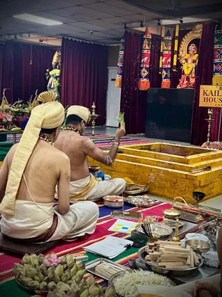 Two priests in white dhotis with cream turbans sit cross-legged on a striped rug in a hall with maroon drapes. The bare-backed priest on the right raises a green leaf toward a yellow-brick homa fire pit, while the priest on the left sits beside him. Brass trays hold flowers, wooden sticks, lotus buds, incense and vessels. Behind them are decorated pillars with colourful cloth, a garlanded deity murti and a shrine figure with yellow and orange garlands. A yellow sign reads KAILASA HOUSTON USA. Most of the scene is a ritual homa setting.