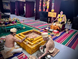 A photo inside a temple hall during a homa fire ritual. Two priests sit bare-chested in white dhotis, one with a white turban and rudraksha-style garland, handling ritual items at a tiered golden yagna kunda with a small flame and a spoon. Behind them on a raised seat under a gold-bordered cloth is a decorated figure of a seated man in a crown and garlands, beside a yellow sign reading KAILASA HOUSTON USA. Striped rugs in pink, green and purple cover the floor; brass oil lamps, offering trays of fruit and sweets, and burnt wood sticks lie nearby. Maroon drapes and colourful banners hang in the background.