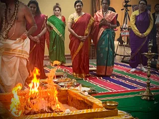 A homa fire burns inside a yellow-banded brick fire pit on a floor covered with striped red, green and blue textile. Behind it, a bare-chested priest in white dhoti and a rudraksha mala stands at the left edge. Four women stand in a row in saris: one in green, one in red with gold border, one in red and green, and one in purple with yellow blouse. A brass oil lamp stands at right and a brass bowl sits near the fire. A camera on a tripod is visible in the background. No one in the scene is identifiable as the a person with appearance-based cues.