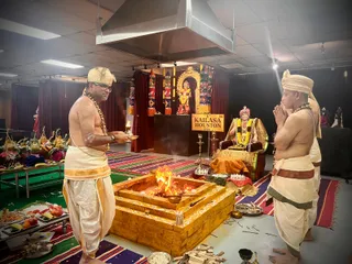 A photograph of a homa ritual in an indoor hall with a KAILASA HOUSTON USA sign on a stand. A square, golden-brick fire pit with a burning fire sits in front of a seated, crowned murti of a figure wearing a gold and red garment, garlanded, on an ornate throne. Behind it stands a deity murti under a decorated arch with red and gold hangings. A priest on the left, bare-chested in cream dhoti with gold border and a gold-trimmed turban, holds a plate and stands near offerings of fruit, flowers and plantains. Two priests on the right, bare-chested with saffron-yellow turbans and garlands, stand with palms pressed together in prayer. Brass lamps and offerings are arranged on the floor and tables.