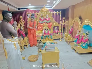 A photograph inside a gold-decorated shrine room during an evening puja. On the left, a shirtless man with a white dhoti edged in yellow stands with palms pressed together in namaskar. Beside him, a woman in an orange saree and rudraksha mala performs a ritual gesture before a golden throne-like altar holding deity murtis garlanded with pink and red flowers. A small brass kalasha, a golden lion-shaped stand, trishula staffs and a golden table with books and a brass plate form the foreground. To the right, a shrine niche holds deity figures in blue and silver garments with flower garlands. A timestamp reads 15 February 2026 at 7:22 pm. No identifiable a person figure is visible.