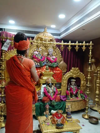 A woman in orange-saffron sari and matching headwrap stands with her back to the camera, hands joined in prayer, before a gold-decorated shrine inside a hall with air-conditioning vents and ceiling lights. The shrine holds a large golden canopy over a pair of silver-crowned deity murtis garlanded with pink and green flowers, a black stone murti of Shiva-like form draped in pink flower garlands and rudraksha, and a smaller murti in a gold arched niche. Brass oil lamps stand on both sides, and a gold table in front holds small figurines, a red cloth bundle, offering bowls and flower petals. A white sign reading KAILASA MADURAI is visible at the left edge.