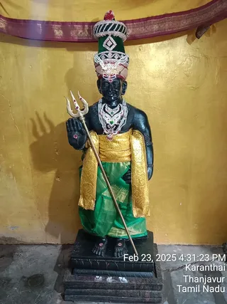 A photograph of a black stone deity murti standing on a dark pedestal against a golden-yellow wall with a maroon and gold bordered cloth canopy above. The figure wears a tall green and silver-jeweled crown with a pink lining, layered silver and jewelled necklaces, a gold waist sash and a green silk dhoti with gold border, and holds a silver trident in its left hand. A camera timestamp in the lower right reads Feb 23, 2025 4:31:23 PM, Karanthai, Thanjavur, Tamil Nadu. No people are visible.