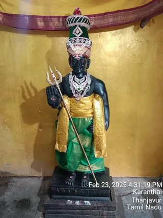 A standing black stone murti of a deity holding a golden trident in its left hand, with its right hand raised in abhaya mudra. The figure wears a tall green and white jeweled crown, layered pearl and ruby necklaces, and a green silk dhoti with gold brocade sashes at the waist and arms. The murti stands on a dark plinth in front of a gold-painted wall draped with a maroon and gold bordered cloth. A date and location timestamp is printed in the lower right corner.