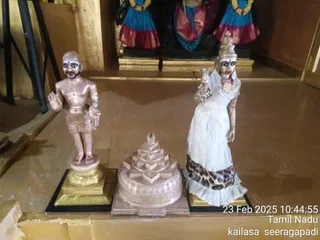 A photograph of three figures on a polished brown floor inside a temple shrine. On the left stands a gold-painted statue of a bare-chested male figure with a red and white tilak on the forehead, wearing a short dhoti and holding one hand raised. In the centre is a tiered pale sri yantra meru structure on a base. On the right stands a gold-painted female figure in a white draped garment with a leopard-print hem, holding a lamp and wearing a red necklace and a gold pendant. Behind them, deity murtis in blue and red silk garments with garlands are visible. A timestamp reads 23 Feb 2025 10:44:55, Tamil Nadu, kailasa seeragapadi.