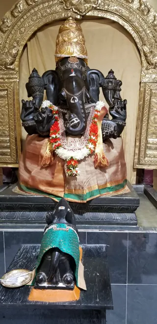 A black stone Ganesha murti sits in a golden arched niche, draped in a copper-and-green silk cloth with a gold border and garlanded with white jasmine and red-orange flowers. A gold crown rests on its head, with a red tilak on the forehead. In front, a smaller black stone figure, a Nandi-like bull or shivalinga-style form, wears a green brocade cloth and a jewelled collar, with a brass plate holding white offerings to its left. No people are visible in the scene.