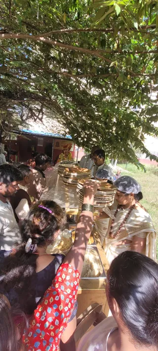Outdoor food distribution (annadan) beneath a large leafy tree in bright daylight. A woman in a white sari with a rudraksha mala and a white head covering stands behind a long table stacked with brass plates and steel serving trays of food. Two men in white clothes stand near her, one wearing a white and gold shawl. Behind the table, a person in a yellow garment with a red turban and flower garland stands near a red-roofed building. Several devotees crowd the table, and a woman in a red patterned top reaches forward with a serving spoon to take food.