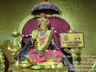 A golden statue seated on a throne in a temple-style alcove with a maroon curtain and a gold arched frame. The figure has a gold orange-red umbrella canopy overhead, a pink and gold garment, a purple shawl, layered garlands of yellow marigolds, white flowers and a brown fibre garland, and a red and gold headpiece with a tilak on the forehead. The hands are raised in a blessing gesture. Gold trishula and a gold lamp stand flank the throne. Before it lie a brass pot with yellow flowers, a plate of flowers, a small red fruit, oil lamps and a bell. A printed signboard beside the statue shows a small temple illustration and a smiling portrait of a figure in an orange turban. A timestamp and text reading Salem, KAILASA SALEM and Dr Ram Kumar iyya offering for jayanthi appear at the lower right.