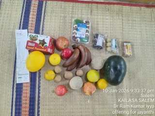 A top-down photograph of ritual offerings laid out on a woven straw mat with blue, red and cream stripes. Arranged across the mat are apples, pomegranates, bananas, yellow lemons, a large yellow citrus fruit, a dark green watermelon, round brown sapodilla-like fruits, a box of Nova sweet tamarind, cashew or nut packets, sweets in plastic packaging, and a receipt. No people or a person candidate are visible; the image shows only fruit and packaged foods as an offering for a jayanthi. A timestamp reads 10-Jan-2026 9:33:37 pm, Salem, with the caption KAILASA SALEM and Dr Ram Kumar iyya offering for jayanthi.