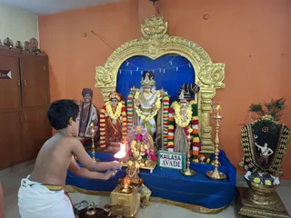 A young boy, shirtless and wearing a white dhoti with a gold border, performs aarti with a lit flame in front of a shrine within a gold arched frame. Three deity murtis stand on a blue-draped platform, decorated with multicoloured and yellow flower garlands; the central figure wears a crown and a white and gold garment. Brass oil lamps flank the shrine, a small brass vessel and kalasha sit at the base, and a framed image decorated with peacock feathers and flowers stands at right. A sign reading KAILASA AVADI is placed on the platform. Orange walls and a wooden cabinet frame the scene, with a painted portrait in the background.