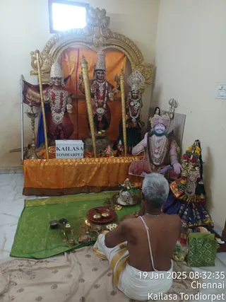 A photograph inside a small room at a KAILASA temple shrine. A seated priest with grey hair, bare-backed and wearing a white dhoti with gold border and a white sacred thread, performs puja on a green cloth in front of a shrine with golden arch. Three deity statues stand in the shrine: left and right figures in red and green clothing with crowns, centre figure holding a trident-like staff and a mace or implement. Two tall gold poles flank them, and an orange cloth hangs behind. To the right stands a large printed cutout of a bearded man in pink-and-red turban with a rudraksha-style garland and a ceremonial gold necklace, seated in a chair beside a decorated purple-clad figure. Brass puja vessels, lamps, a kalasha, flowers and a red plate sit on the green cloth. Wall text on a white box reads KAILASA TONDIARPET.