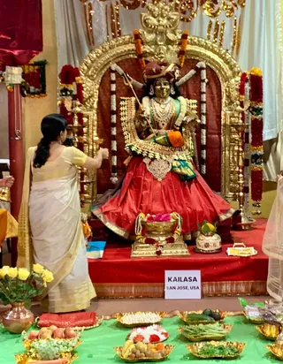 A photograph of a large gilded deity murti seated on a red-draped platform, decorated with marigold and jasmine garlands and a red-and-gold skirt, holding a trident-like weapon, with a gold arched backdrop. In front stands a woman in a cream and gold-bordered sari with her back to the camera, raising her hands near a brass oil lamp. Before the murti are a golden kalasha-style vessel garlanded with flowers, and brass plates of fruit, green vegetables and sweets on a green cloth. A small white sign reads KAILASA SAN JOSE, USA. No a person candidate is visible.