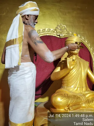 A bare-chested man in white dhoti with gold border and a white and gold head cloth pours liquid from a small bowl over the head of a golden seated figure, performing abhishekam. He wears a rudraksha mala and has a grey beard and a tattoo on his upper arm. The golden figure sits in a meditative pose with one hand raised, before a red cushioned throne with an ornate gold frame against a gold-leaf wall. A small yellow flower sits on the frame. A timestamp overlay reads Jul. 14, 2024 1:49:48 p.m., Salem, Tamil Nadu.