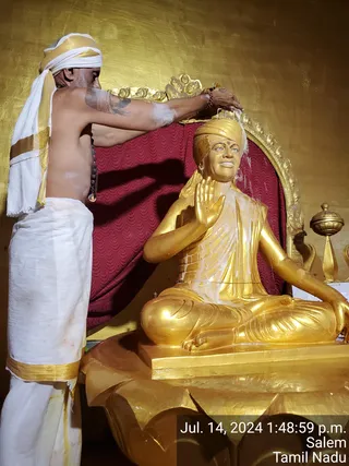 A shirtless priest in a white dhoti with gold border and white-and-gold head cloth pours liquid over a seated golden statue. The statue, with a golden turban and raised right hand in blessing, sits in lotus position on a golden lotus base before a red draped backdrop and gilded throne. The priest wears a rudraksha mala and has white sacred ash marks on his body while performing abhishekam. A timestamp reads Jul. 14, 2024, Salem, Tamil Nadu.