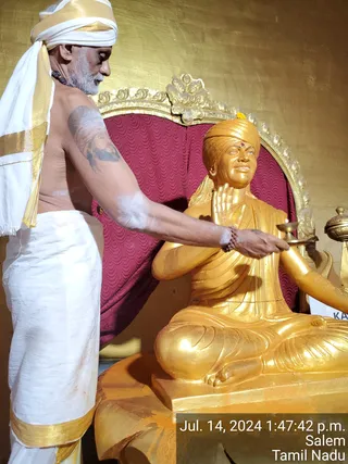 A shirtless, grey-bearded man in a white dhoti with gold border and a white shawl with gold edging stands at left, holding a small oil lamp toward a golden statue. The statue is a seated figure in a golden turban with a red dot on the forehead, draped in a golden robe, set before a deep red patterned cushioned chair with an ornate gold frame. The man wears a rudraksha mala on his wrist and has a tattoo of a bearded face on his upper arm. The scene shows a ritual offering of light to a golden murti in a gold-walled shrine, with a partial 'KA' sign visible at right and a timestamp reading Jul. 14, 2024, Salem, Tamil Nadu.