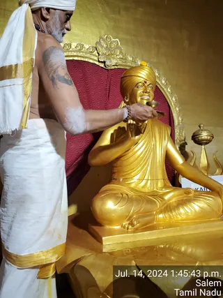 A photograph of a priest performing abhishekam on a golden seated statue. The priest stands at left, shirtless with a white head cloth and white dhoti bordered in gold, a sacred ash mark on his arm and a rudraksha bead bracelet on his wrist. He holds a small cup or vessel up to the statue's chest. The statue is a gold-painted seated figure in a yellow turban with a red forehead mark, wrapped in a flowing robe, set before a red patterned backdrop inside an ornate gold frame. A gold trident-topped ritual object stands at right. A timestamp overlay reads Jul. 14, 2024 1:45:43 p.m., Salem, Tamil Nadu.