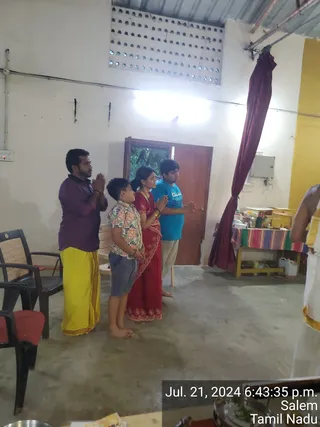 Photograph of a family standing with palms pressed together in namaskar in a plain concrete hall. A man in a purple shirt and yellow dhoti stands at left, next to a boy in a floral shirt and grey shorts, then a woman in a red saree with gold border and a boy in a blue t-shirt. A dark red curtain hangs from the ceiling at right beside a table with a striped cloth and puja items. A partially visible person in white dhoti with gold border stands at the right edge. A timestamp reads Jul. 21, 2024 6:43:35 p.m. Salem Tamil Nadu. Four people are visible.