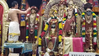 A shirtless priest in white dhoti with a yellow border stands at left, holding a small ritual vessel and offering to a grouping of gilded metal murtis on a decorated altar. Central figure is a dark stone deity murti with a turban-style crown, adorned with fresh garlands of yellow, red and white flowers and rudraksha beads. Flanking it are two gilded standing figures: a male figure in saffron-brown robes with a red-and-white turban and layered gold necklaces, and a female figure with an ornate crown and dark garment. A bearded figure in brown robes and turban stands at right, also garlanded. Brass oil lamps, bells and bowls sit on a blue-cloth table and the altar below. A pink cloth and a white placard reading KAILASA LOS ANGELES are visible at bottom right. Colourful painted panels frame the scene.