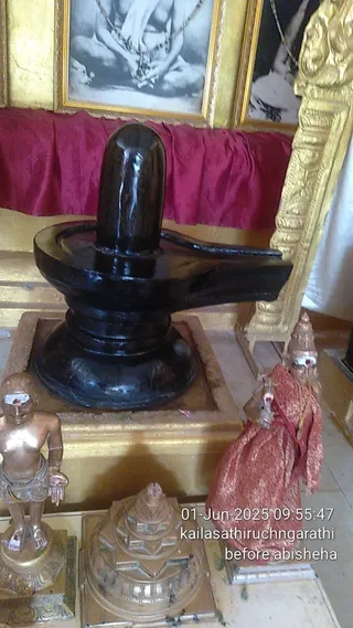 A dark polished Shiva lingam on a black yoni-pitha base stands on a gold-framed altar platform before a red draped cloth. Behind it, two framed black-and-white photographs hang in gold frames. To the left, a small bronze standing figure with white ash markings and a tilak on its forehead stands on a pedestal. In the lower foreground, a tiered bronze pyramid-shaped object sits on the floor. To the right, a figure wrapped in a red-and-orange patterned cloth with a red sash stands beside a gold ornate frame. No physical people are visible. Timestamp overlay reads 01-Jun-2025 09:55:47.