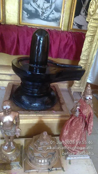 A dark polished black stone Shivalinga stands on a square gold-trimmed base inside a shrine before abhishekam. Behind it hang a deep red silk drape and gilded frames holding black-and-white portraits. To the lower left a small bronze standing figure with a white tilak and loincloth, and a small bronze pyramidal shrine model sit on the floor. To the right a bronze deity figure wrapped in a red-orange cloth stands beside the linga. No people are visible. A timestamp reads 01-Jun-2025 09:55:46 with a caption about the ritual before abhisheha.