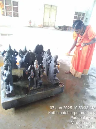 A woman in an orange dress with a gold-bordered hem stands at the right, holding a brass plate and a small vessel while bending toward a black stone shrine. On the shrine stands a dark stone deity murti with a crown, raised arms and decorated pillars behind it, with a small bowl of rudraksha beads placed at its base. The floor is wet and the hall is white-walled with barred windows and a double door. A timestamp overlay reads 07-Jun-2025 10:53:33 AM, Kothainatchiarpuram Road, Pudupalaiyam, Virudhunagar, Tamil Nadu. No a person candidate is visible.