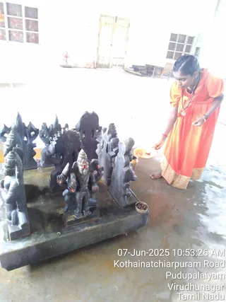 A woman in an orange dress with gold-edged hem stands barefoot at the right, bending toward a brass plate of offerings on a tiled floor, with a rudraksha mala around her neck. In front of her on a dark stone platform is a set of dark carved stone sculptures, including a central standing deity murti with a conical crown and multiple arms, flanked by carved panels and figures. A small bowl of rudraksha beads sits at the platform's edge. The room is plain white with windows, a doorway and a small figure in the background. A timestamp reads 07-Jun-2025 and a Tamil Nadu location overlay appears in the corner.