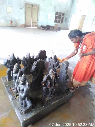 A photograph of a woman in an orange dress with gold-bordered hem performing puja at a black stone sculptural arrangement of deity murtis on a raised black stone platform. She wears a rudraksha-style mala and a small round pendant, holds a small bell in one hand and a brass plate in the other, and bends toward the murtis. A small bowl of offerings sits at the platform's edge. Behind them is a bare hall with pale green walls, double wooden doors, a window and a small standing figure on a stand. A timestamp in the lower right reads 07-Jun-2025 10:52:38 AM.