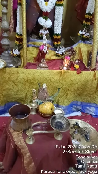 A photograph of a small temple shrine draped in gold and maroon cloth, with white and yellow flower garlands hanging on the sides. At the centre stands a small metal deity murti with a red cloth and a silver-trimmed crown, flanked by a lit oil lamp. In the foreground are brass vessels: a copper pot, a brass lamp stand, a round yellow offering, and a brass plate with small items. No people are visible. A timestamp and location caption in the lower right read 15 Jun 2025, Tondiarpet, Chennai, Tamil Nadu.
