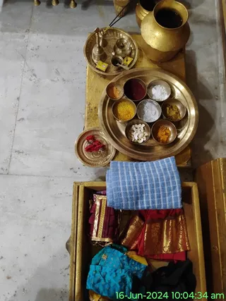 A photograph taken from above of a temple ritual preparation setup on a wooden stand over a grey stone floor. A large brass plate holds small bowls of coloured powders in orange, red, yellow and white, plus cashew nuts and a yellow turmeric-like powder. Beside it is a brass tray with a bell, a small oil lamp and a brass kalasha-style pot with dark liquid. A smaller brass plate holds red flowers. Below, an open wooden box contains folded cloths including a blue checked towel, a maroon garment with gold border, a red and gold silk cloth and a turquoise embroidered garment. A timestamp reads 16-Jun-2024 10:04:34 am. No people are visible.