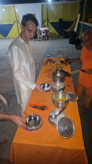 Photo of an outdoor evening food service at night. A young man in a cream-coloured long kurta with vertical stripes stands at a long table draped in orange cloth, holding a small stainless steel bowl. The table holds several large stainless steel pots with lids, bowls of food, spoons on an orange tray, and a pink-rimmed plate. On the right, a blurred man in an orange top and orange cloth pours or serves from the table. In the background, a seated man in white sits on a red chair near a yellow and blue wall with a white cloth hung above. a person is not visibly identifiable here.