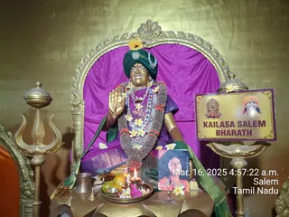 A golden statue of a seated figure in a shrine, dressed in a green turban with a yellow flower on top, a purple dress and a green shawl, draped with garlands of white and yellow flowers and a thick brown-and-red garland of leaves. The statue's right hand is raised in blessing. In front is a brass plate with a lit oil lamp, orange fruits, flowers, a small brass cup and a printed booklet. Brass trishula-topped stands flank the golden arch behind, and a yellow board reads KAILASA SALEM BHARATH beside small pictures. The statue is a depiction, not a living person, so no physical people are present.