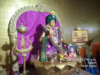 A golden statue of a seated figure in a green turban with a gold brooch and yellow hibiscus, wearing a purple garment, green sash and layered flower and marigold garlands, hands pressed together in namaskar. It sits on a golden lotus-shaped pedestal before a magenta drape and an ornate gold arched frame. Offerings on the table include a lit oil lamp, a brass bell, a small kalasha-like vessel, bananas, mangoes, flowers and a framed portrait. A gold trident stands at left, and a placard at right reads KAILASA SALEM BHARATH beside a small portrait. The photo carries a timestamp of Mar. 16, 2025, 4:57:49 a.m., Salem, Tamil Nadu.