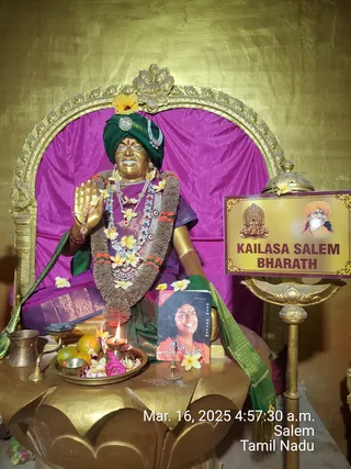 A golden statue seated on a purple cushioned throne with an ornate gold frame. The figure wears a green turban with a brooch, layered pearl and jewelled necklaces, a flower garland, and a purple garment with a green shawl; its right hand is raised in blessing. Yellow plumeria flowers are scattered around it. In front, a brass plate holds oranges, flowers and a lit oil lamp, beside a brass pot and a small book with a portrait of a smiling woman. A gold sign reads KAILASA SALEM BHARATH and shows a temple and a bearded figure in an orange turban. A timestamp reads Mar. 16, 2025 4:57:30 a.m. Salem, Tamil Nadu. No people are visible.
