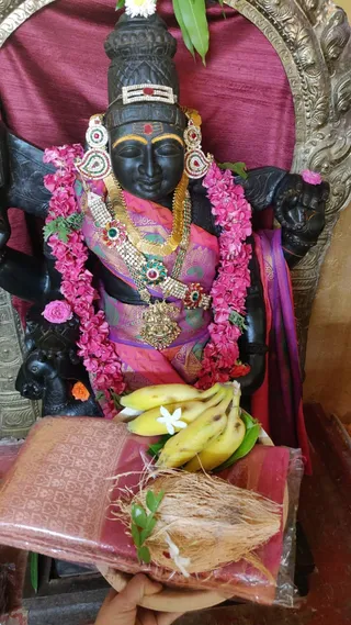 A dark stone murti of a deity stands in a shrine, adorned with pink and magenta flower garlands, gold and jewelled necklaces, a red-and-gold tilak, and a gold-trimmed crown. The murti wears a pink and purple silk garment and holds a flower in one raised hand. In front, offerings are laid out: bananas, a coconut, a folded red-and-gold silk cloth, and a white flower. A hand at the bottom edge holds the offering plate. A purple cloth drapes over the murti's arm, and a maroon backdrop frames the shrine.