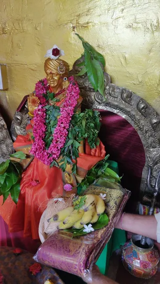 A golden statue of a seated figure in an orange-red sari with gold border and a golden turban, hands joined in namaskar, stands in a shrine before a gold-leafed wall. The statue wears a pink flower garland with green leaves, a gold necklace and a red-and-white flower on its head. Behind it is an ornate silver-toned throne back with a maroon cushion and a mango leaf toran. In front are a brass-bordered tray with bananas, a coconut, jasmine flowers and a green cloth, plus a decorated brass pot and a hand of a devotee at the edge. No living people are clearly visible apart from that hand.