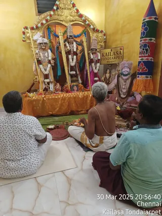 A photograph inside a gold-framed shrine at KAILASA Tondaiarpet, Chennai. Three male devotees sit on the floor facing the shrine: the left man wears a white patterned shirt and white dhoti, the right man wears a light green shirt and maroon cloth, and a shaved-headed priest in a bare-chested pose with a white sacred thread and white dhoti performs a puja with a brass plate of offerings. Three deity statues stand under a gold arch decorated with red and white flower garlands; the central figure wears a tall crown and a garland, the left figure a white turban, and the right figure a pointed crown. A standing statue of a figure with pink turban, rudraksha mala and ornaments stands at the right of the shrine. A signboard reads KAILASA TONDIARPET BHARAT, and a small framed portrait is placed on the orange cloth-draped altar, with a banner of blue and red floral design at the right. A timestamp reads 30 Mar 2025 20:56:40 Chennai.