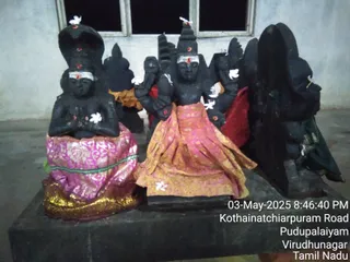 A photograph of a group of dark stone murti statues on a raised black stone platform, lit at night. The central figure has four arms and a crown, with white tilak and red dot markings on its forehead. A seated figure on the left, with hands joined in namaskar and a hooded serpent above its head, wears a pink and gold brocade cloth. The central statue is draped in an orange and gold patterned cloth. White flowers are placed on the statues, and an orange cloth hangs behind them. On the right, a tall dark stone figure wears a green cloth. Behind them, a window and plain whitewashed wall. A timestamp and location caption are printed on the lower right: Kothainatchiarpuram Road, Pudupalaiyam, Virudhunagar, Tamil Nadu.