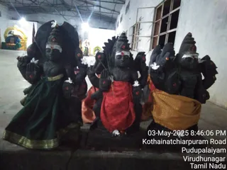 Photograph taken at night inside a plain whitewashed hall of a temple, showing three dark black stone murti figures in a row, each with multiple arms, white tilaka, red kumkum dots and white flowers placed on them. The left figure wears a green skirt with gold border, the centre figure a red cloth, and the right figure a golden-yellow cloth. In the background a gilded throne-like seat with a blue cushion is visible, and a bright overhead light illuminates the hall. A date stamp reads 03-May-2025 8:46:06 PM Kothainatchiarpuram Road, Pudupalaiyam, Virudhunagar, Tamil Nadu.